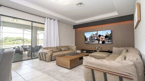Ein modernes Wohnzimmer mit einem großen Flachbildfernseher, einer Sofagarnitur, einem Couchtisch und einer Glasschiebetür zu einem Außenbereich.