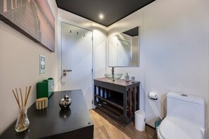 Une salle de bain moderne avec un lavabo en forme de bol en verre, un miroir, des toilettes et une petite table sur laquelle se trouvent un vase et une bouteille.