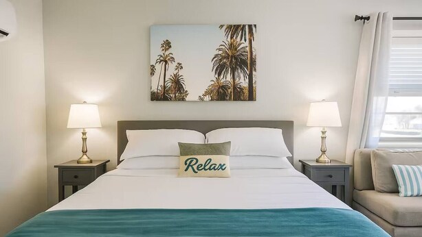Een hotelkamer met een bed, twee bedlampen, een kussen met de tekst 'Relax', een schilderij van palmbomen en een raam met gordijnen.