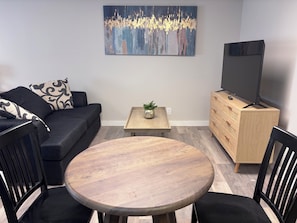 A living room with a sofa, a coffee table, a TV on a stand, and a dining table with chairs.