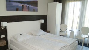 Habitación de hotel con cama, dos sillas, una mesita, un cuadro en la pared y un armario blanco grande.