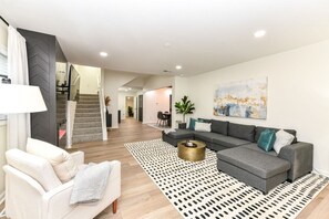 Una sala moderna con un sofá seccional gris, un sillón blanco, un tapete estampado y una escalera que lleva a otro nivel.