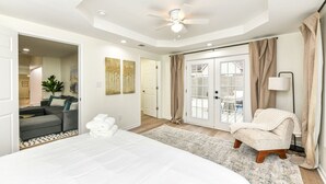 A spacious bedroom with a large bed, a sitting area with a sofa, a chair, and a view of the backyard through glass doors.