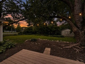 A wooden deck, a tree, and a shed in a backyard at dusk. - Grand Park | King Suite | Fenced Yard | Sleeps 8 (Westfield)