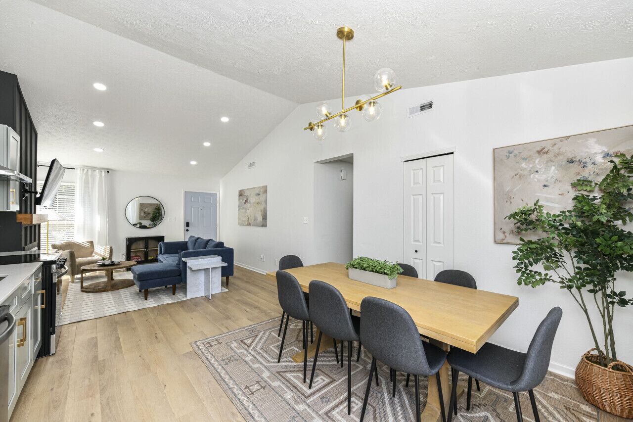 A modern kitchen with a built-in oven, a dining area with a wooden table and grey chairs, and a living room with a blue sofa.