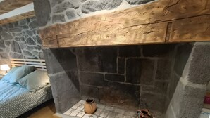 En eldstad i sten med två vaser, en säng med mönstrat sänglinne och en lampa på ett nattduksbord.