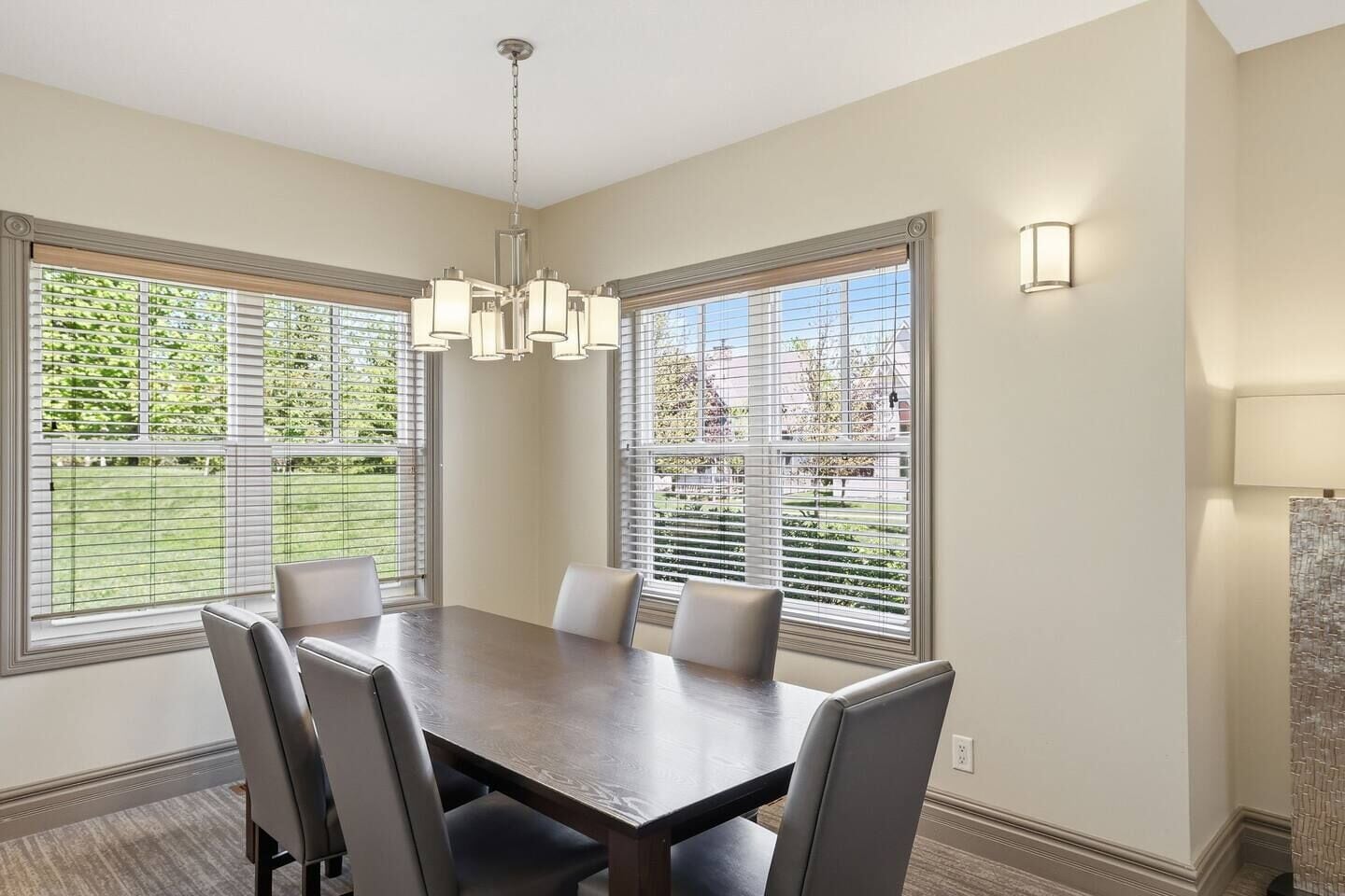 A dining room with a large table, chairs, a chandelier, and windows with blinds.