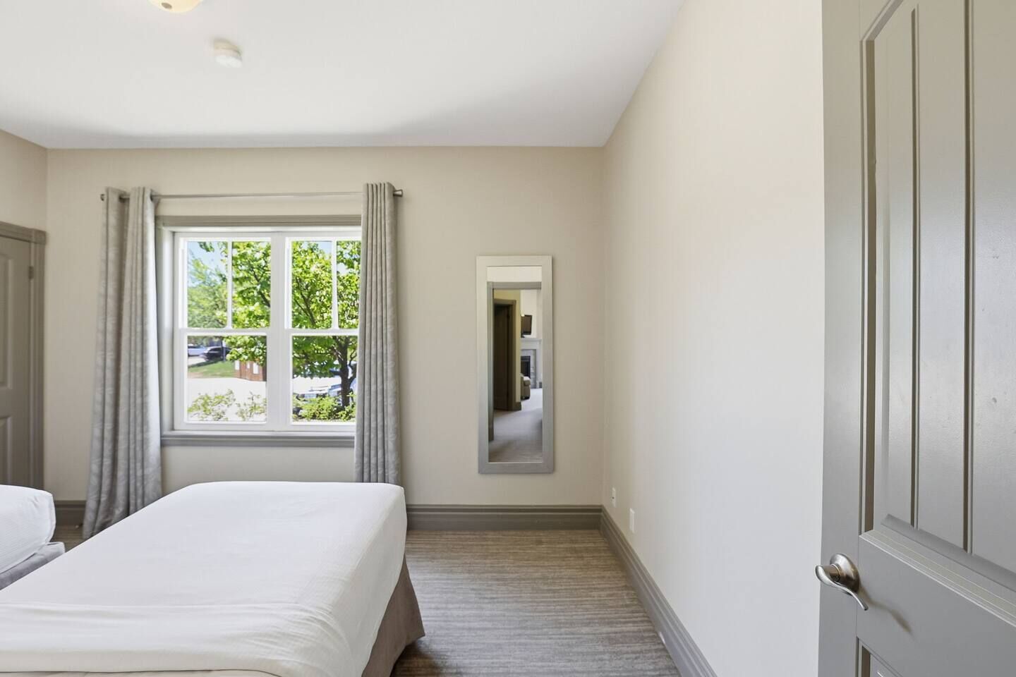 A hotel room with a bed, a window with curtains, a mirror, and a door.