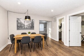 A dining area with a wooden table and chairs, a black pendant light, and a black and white cityscape painting on the wall. - Grant’s Fresh Modern Charm 3B 2B (Noblesville)