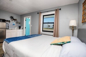 Ein Hotelzimmer mit Bett, Schreibtisch mit Stuhl, Fenster mit Vorhängen, Lampe und Blick ins Freie.