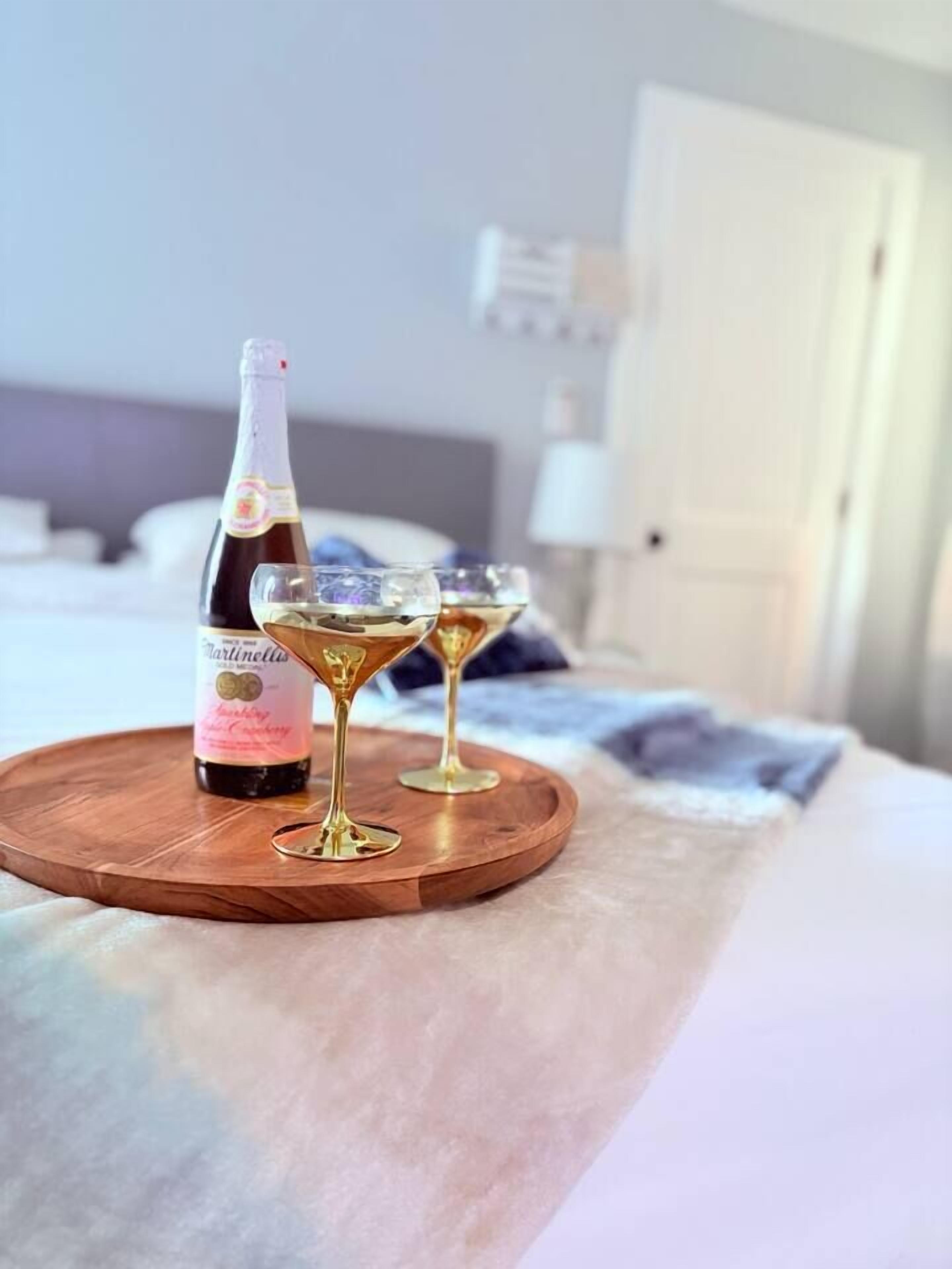 Une bouteille de liqueur Martini et deux verres sur un plateau en bois, posés sur un lit.