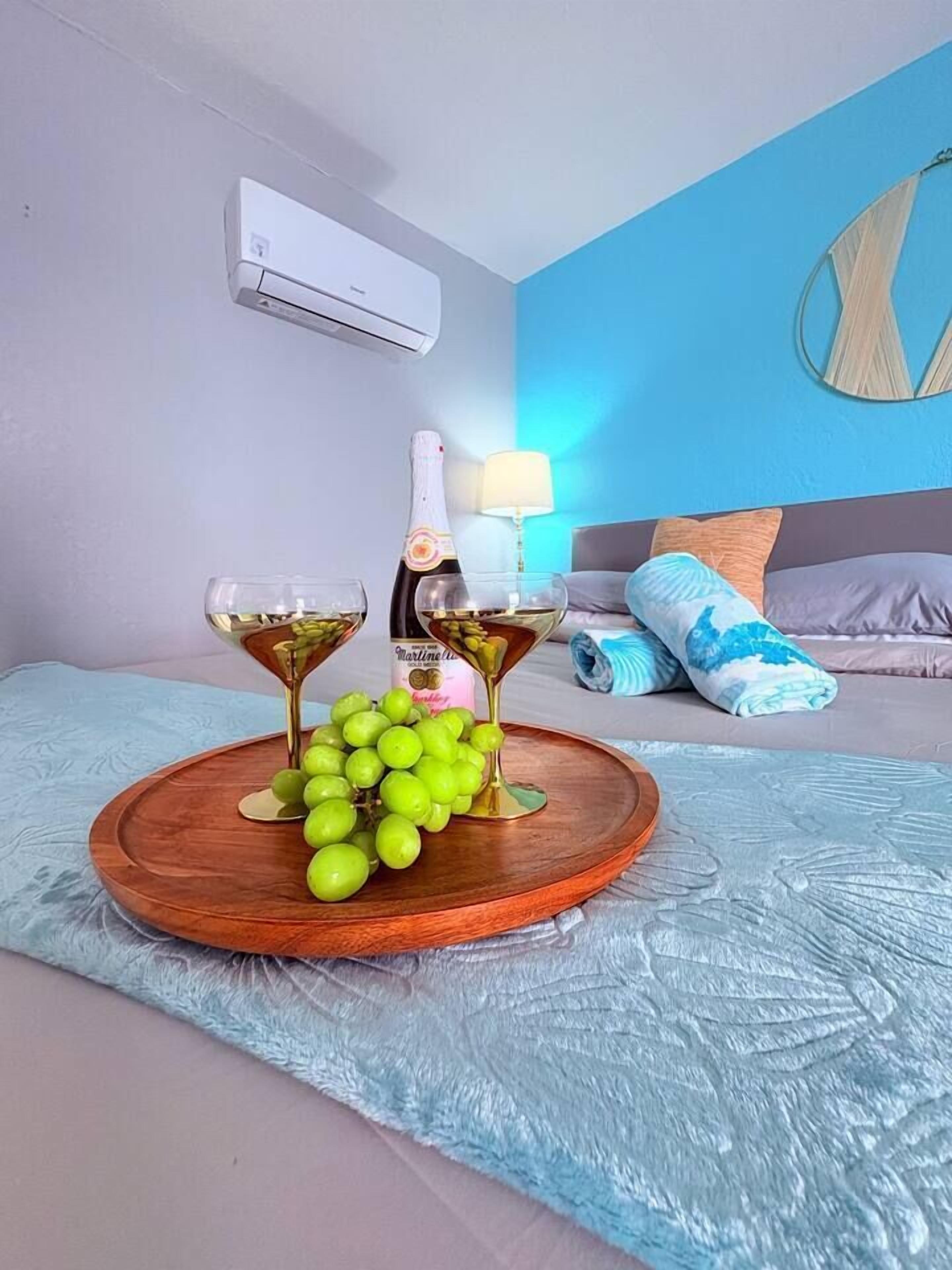 A hotel room with a bed, a bedside table with a bottle and glasses, a wall-mounted air conditioner, and a blue wall.