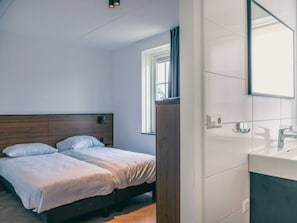 Ein Schlafzimmer mit Holzkopfstütze, zwei Betten, einem Fenster mit Vorhängen sowie einem Badezimmer mit Spiegel und Waschbecken.