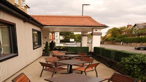 Eine Terrasse im Freien mit Tischen und Stühlen, ein überdachter Sitzbereich und ein Blick auf die Straße und die Gebäude im Hintergrund.