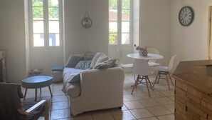 Een kamer met een witte bank, een ronde tafel met stoelen, een klok aan de muur en grote ramen.