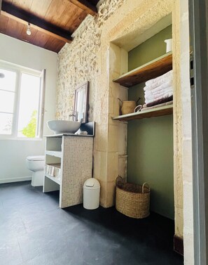 A bathroom with a stone wall, a sink, a toilet, and shelves with towels. - "At the Sundial" Cottage on the banks of the Charente (Port-d'Envaux)