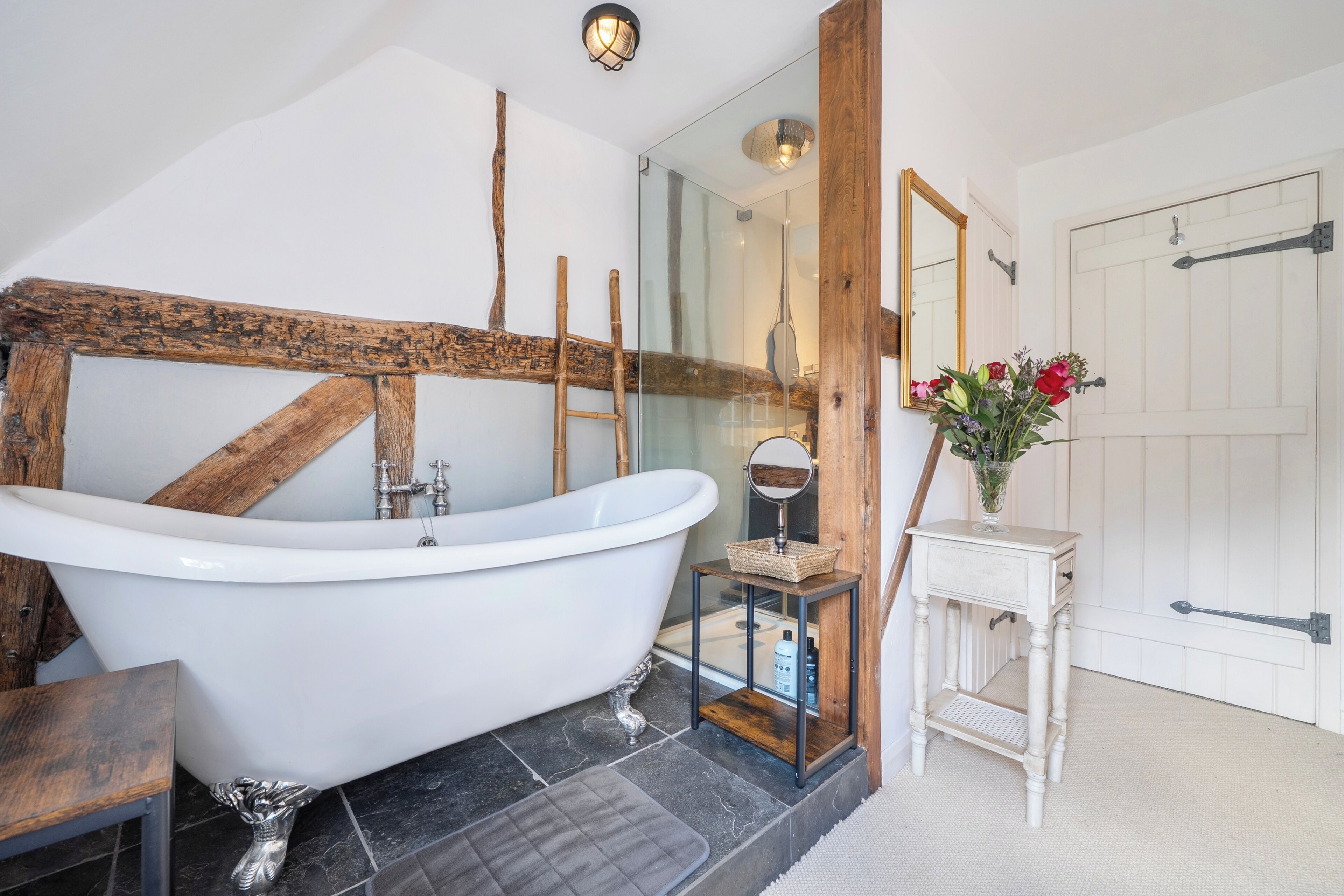 A bathroom with a freestanding bathtub, a wooden beam, a small table with a vase, and a door with a handle.