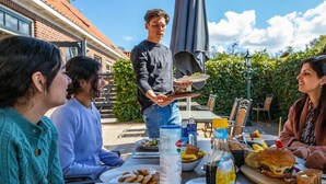 An outdoor dining area with a table set for a meal, a person serving food, and a brick building in the background. - Holiday Home in Velsen-zuid With Terrace (Velsen-Zuid)