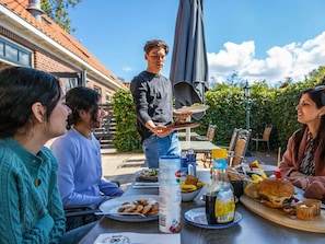 Ein Essbereich im Freien mit einem für eine Mahlzeit gedeckten Tisch, einer Person, die Speisen serviert, und einem Backsteinhaus im Hintergrund.