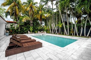 Piscina com deck ladrilhado, cercada por palmeiras e um edifício branco.