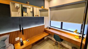 Een moderne kamer met een houten bankje, een bureau, een stoel, een groot raam met een zonnescherm en een spiegel aan de muur.