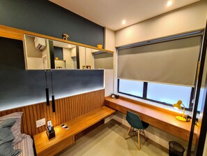 Een moderne kamer met een houten bankje, een bureau, een stoel, een groot raam met een zonnescherm en een spiegel aan de muur.