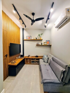 A modern living room with a flat-screen TV, wooden panel wall, ceiling fan, and built-in shelves. - Studio do Arquiteto 1 (Rio de Janeiro)