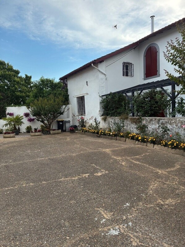 A white building with a red door and windows, surrounded by a gravel driveway and flower beds. - Apartment for rent in the Basque Country near the beaches of Biarritz and the mountains (USTARITZ)