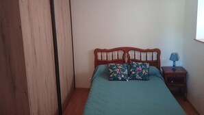 A bedroom with a wooden bed frame, two floral pillows, a blue bedspread, a wooden nightstand with a lamp, and a wardrobe with a black trim. - Apartment for rent in the Basque Country near the beaches of Biarritz and the mountains (USTARITZ)