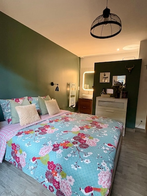 Ein Schlafzimmer mit floralem Bettbezug, einer Pendelleuchte, einem Spiegel und einer Kommode.