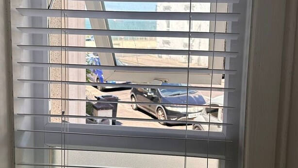 A window with horizontal blinds, a view of a street with parked cars, and a building in the background.