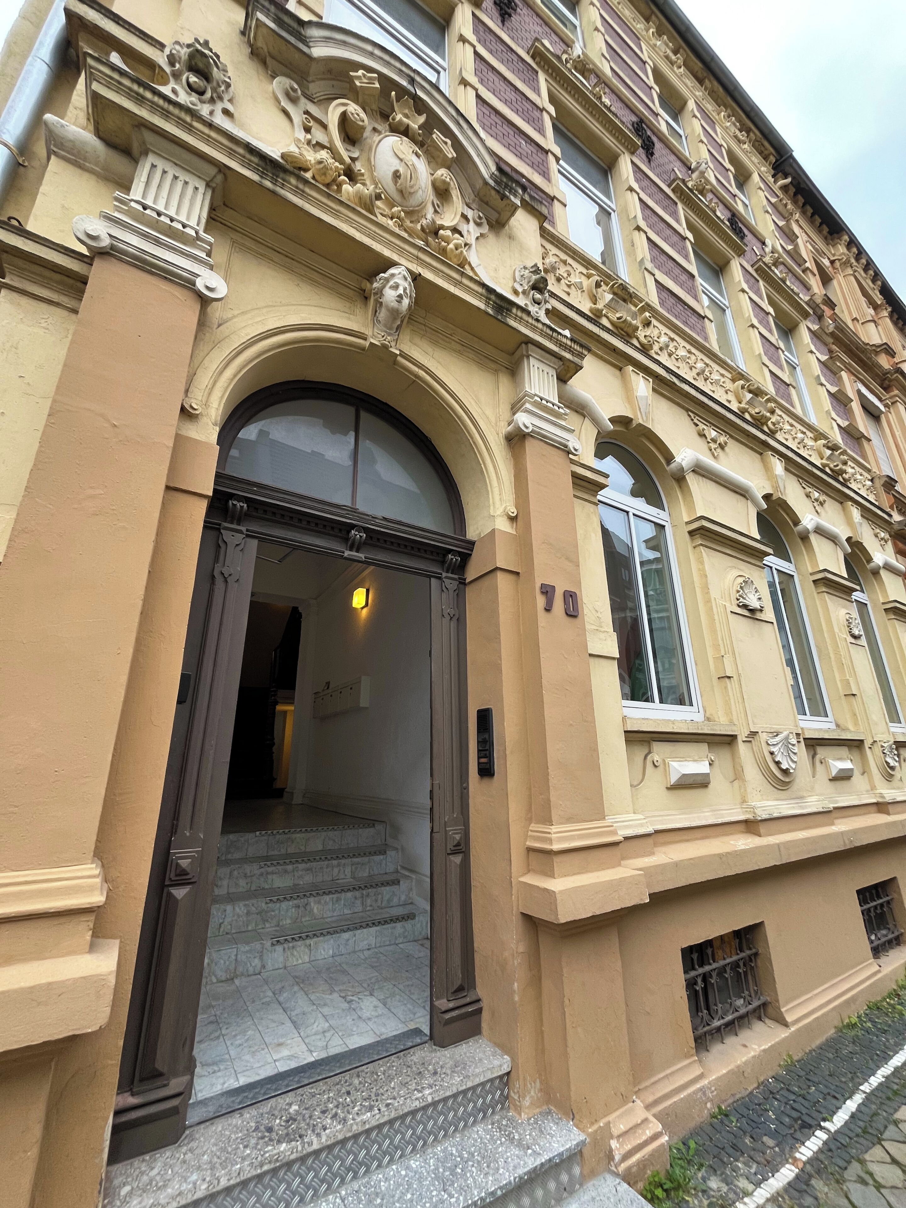 Ein Gebäude mit einem großen Bogenportal, einer Treppe, die zum Eingang führt, und der Hausnummer 70 an der Fassade.