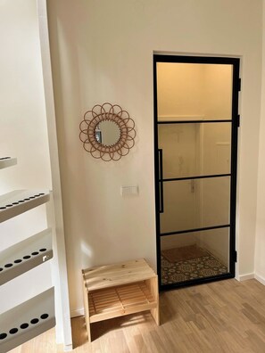 Ein Zimmer mit einer hölzernen Bank, einem an der Wand befestigten Spiegel und einer Schiebeglastür zum Badezimmer.