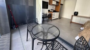 A modern room with a glass-top table, black metal chairs, a bed, a kitchen area, and a red broom leaning against the wall. - Estúdio com piscina e academia no rooftop (São Paulo)