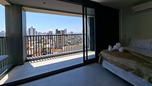A balcony with a city view, a bed with pillows, and a window with blinds. - Estúdio com piscina e academia no rooftop (São Paulo)