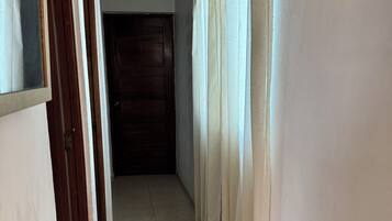 Un pasillo con una puerta de madera al final, paredes blancas y suelo de baldosas.