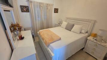 Un dormitorio con cama, mesitas de noche, cómoda, espejo y ventana con cortinas.