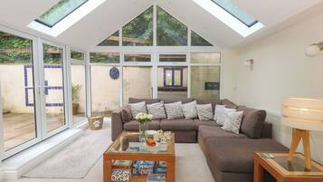 A modern living room with a high ceiling, large windows, a brown sofa, a coffee table with books, and a lamp.