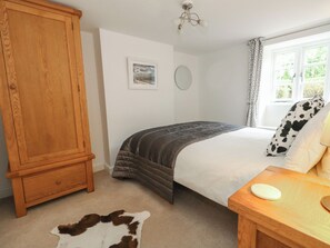A bedroom with a bed, a wooden wardrobe, a mirror, and a window with curtains. - Woodbine Cottage (Weymouth)