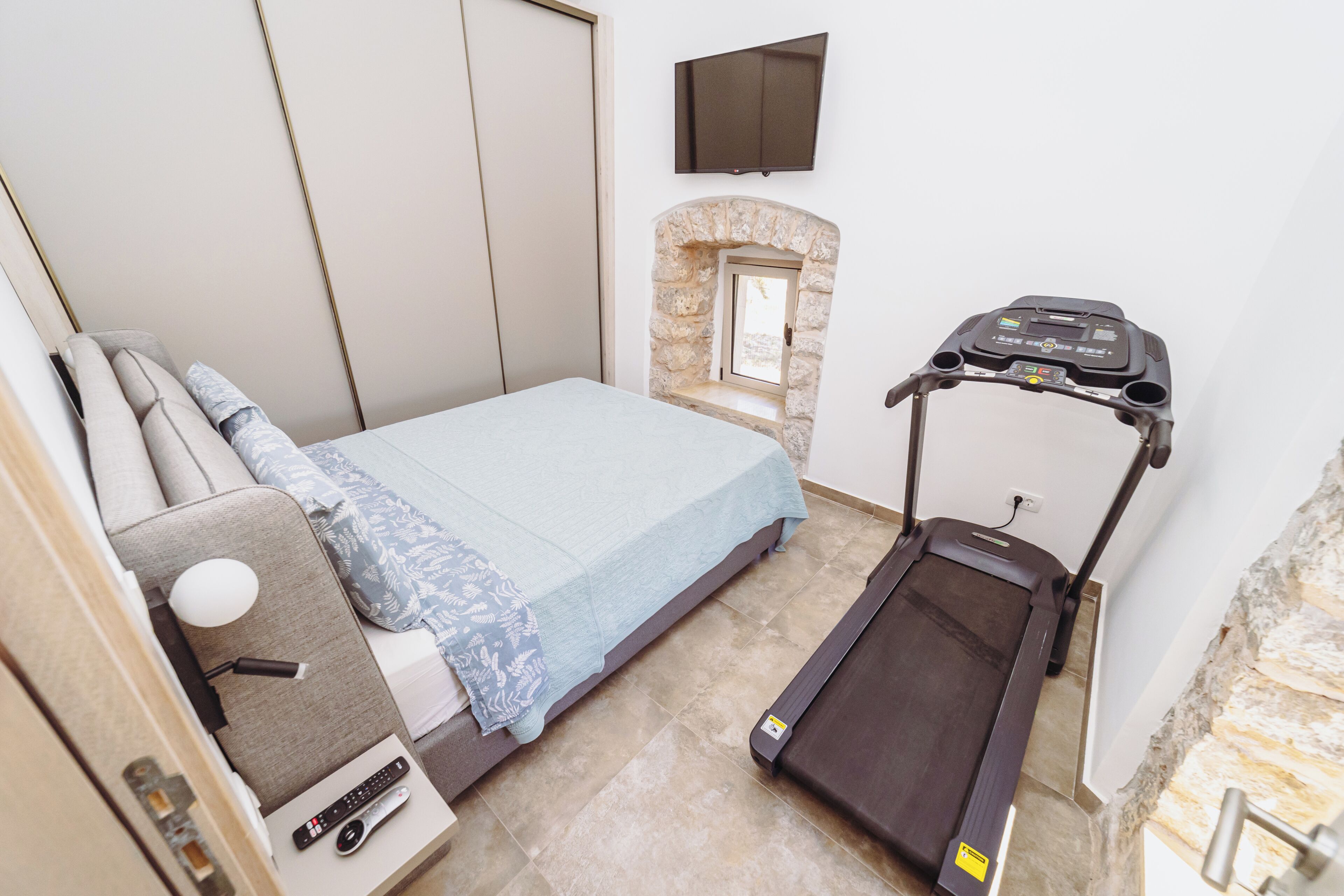 A bedroom with a bed, a treadmill, a TV, and a stone fireplace.