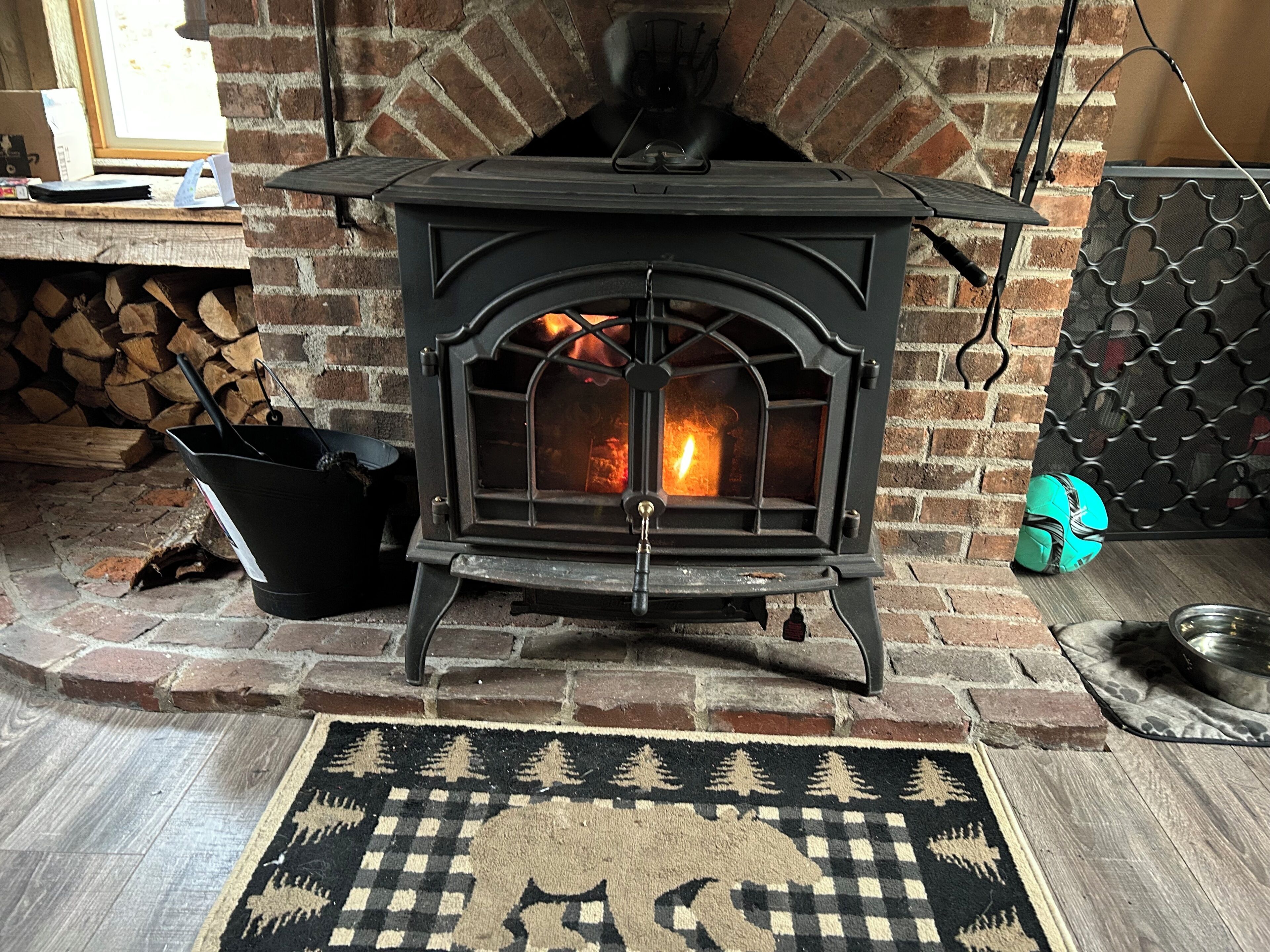A wood-burning stove with a brick wall behind it, a rug with a forest pattern, and a metal basket.