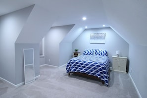 A bedroom with a sloped ceiling, a bed with a patterned bedspread, a nightstand with a lamp, and a small window.