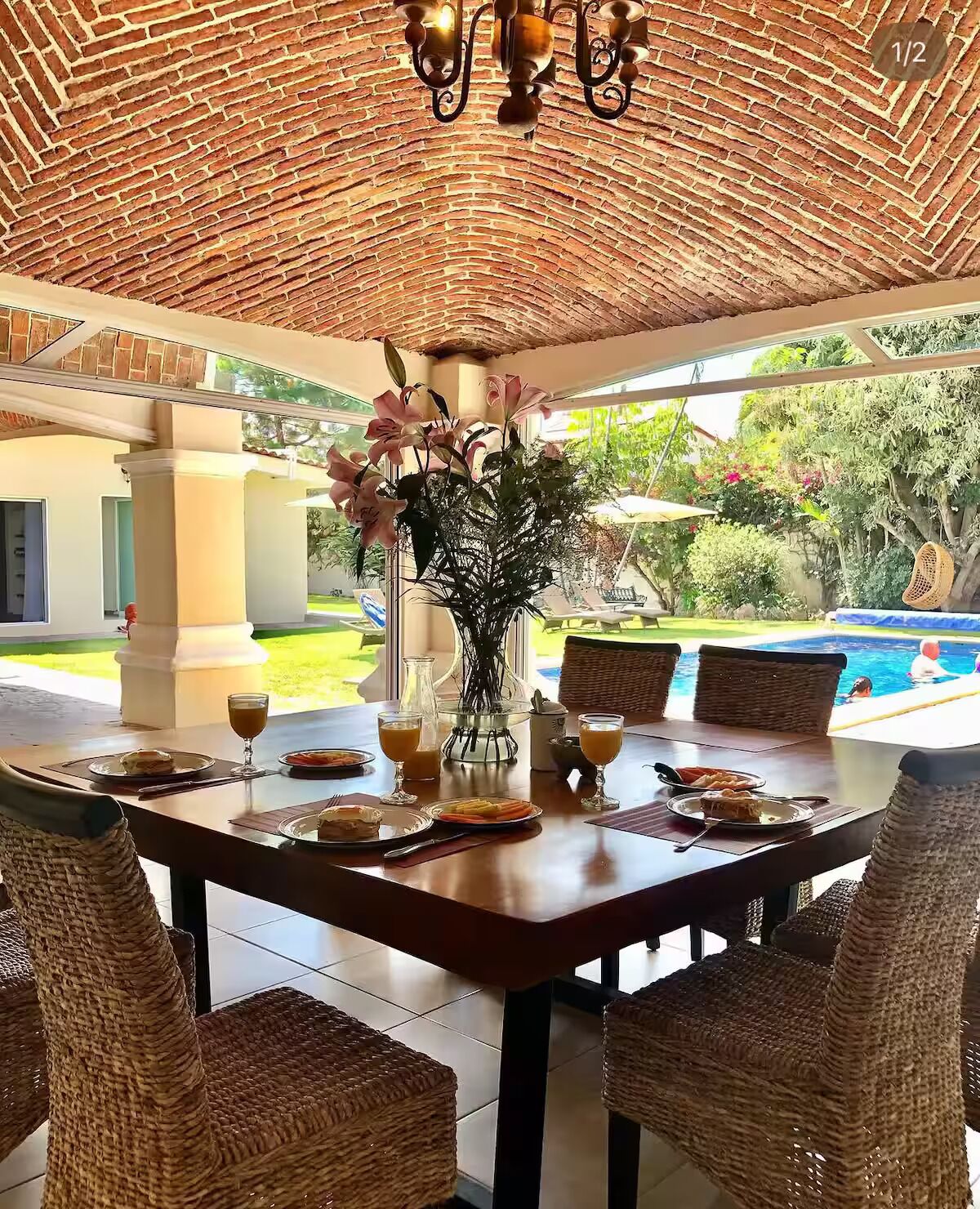 Un espace extérieur abrité, aménagé pour les repas, avec des chaises en osier, une table dressée pour le petit-déjeuner et une vue sur une piscine.