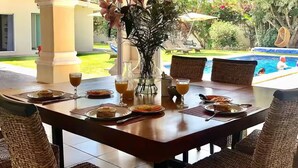 A covered outdoor dining area with wicker chairs, a table set for breakfast, and a view of a swimming pool.