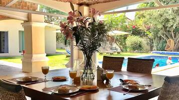 Un espace extérieur abrité, aménagé pour les repas, avec des chaises en osier, une table dressée pour le petit-déjeuner et une vue sur une piscine.