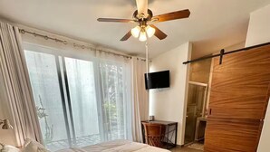 A bedroom with a bed, a desk, a chair, a ceiling fan, a TV, and a sliding door.