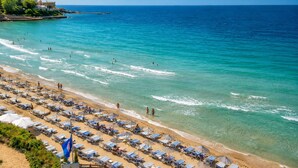 Una spiaggia con file di lettini e ombrelloni, un mare blu limpido e edifici sullo sfondo.