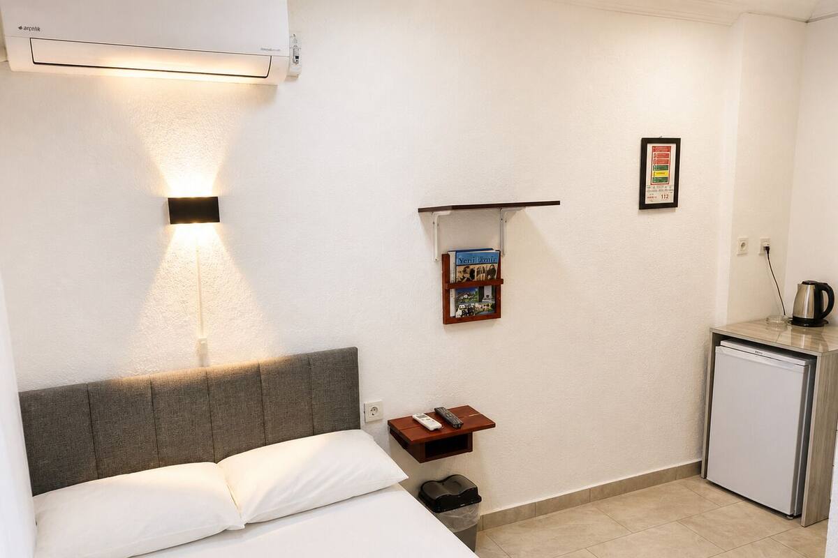 Habitació d'hotel amb un llit, una cuineta petita, un gresol, una prestatgeria amb un llibre i un aire condicionat fixat a la paret.