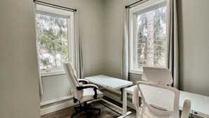 Ein Zimmer mit zwei weißen Schreibtischen, einem weißen Bürostuhl und einem rollbaren Stuhl. Es gibt Fenster mit Vorhängen, eine Deckenleuchte und einen Holzboden.