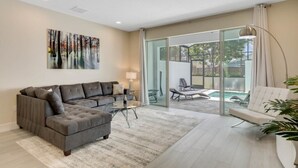 Ein modernes Wohnzimmer mit einem Ecksofa, einer Glastür, die auf einen Balkon führt, und Blick auf einen Pool.
